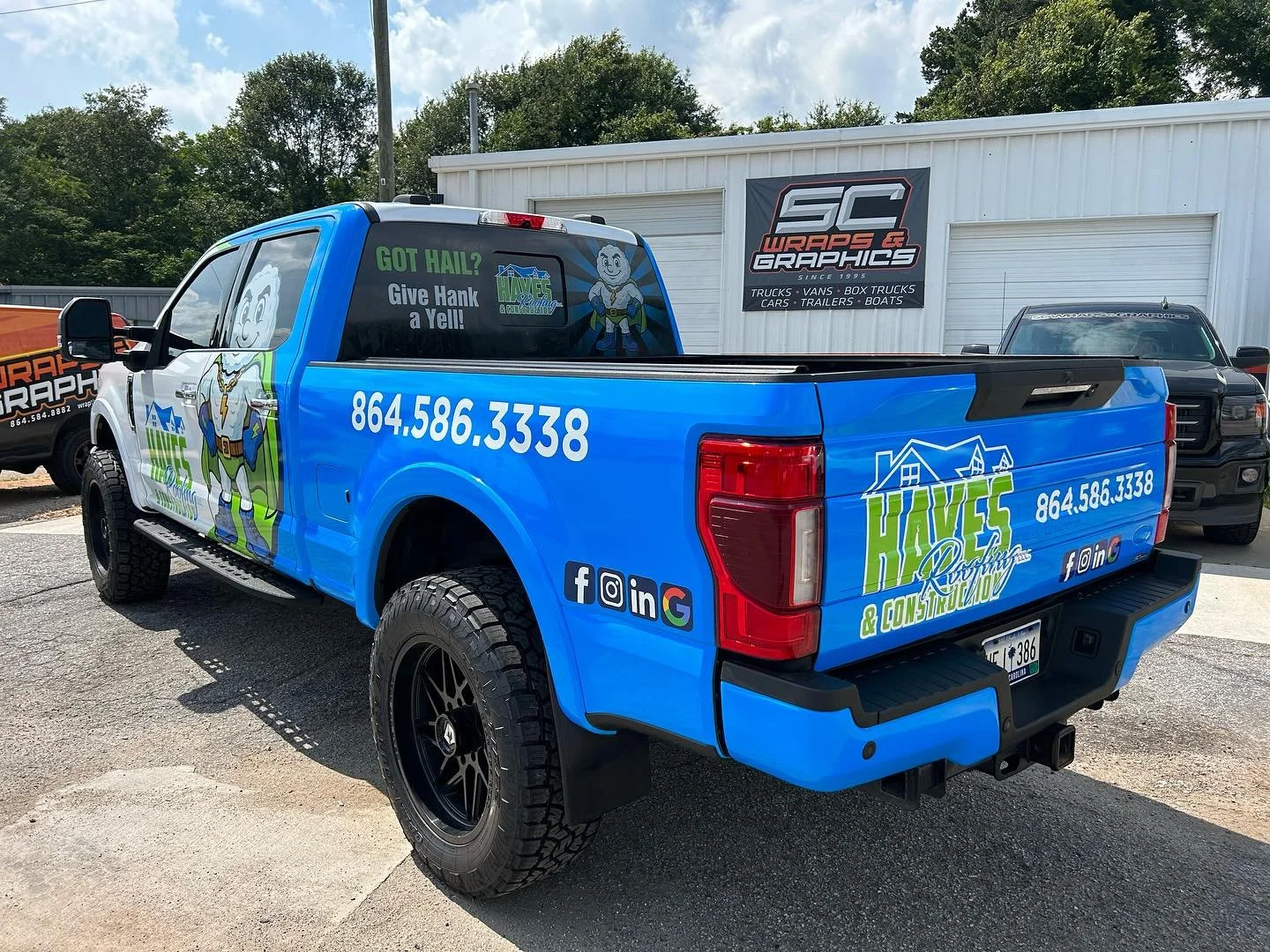 Blue pickup truck with advertising for Hives Roofing & Construction, featuring a cartoon beekeeper logo, phone number 864.586.3338, and social media icons, parked outside a building with a sign reading 'SC Wraps & Graphics'.