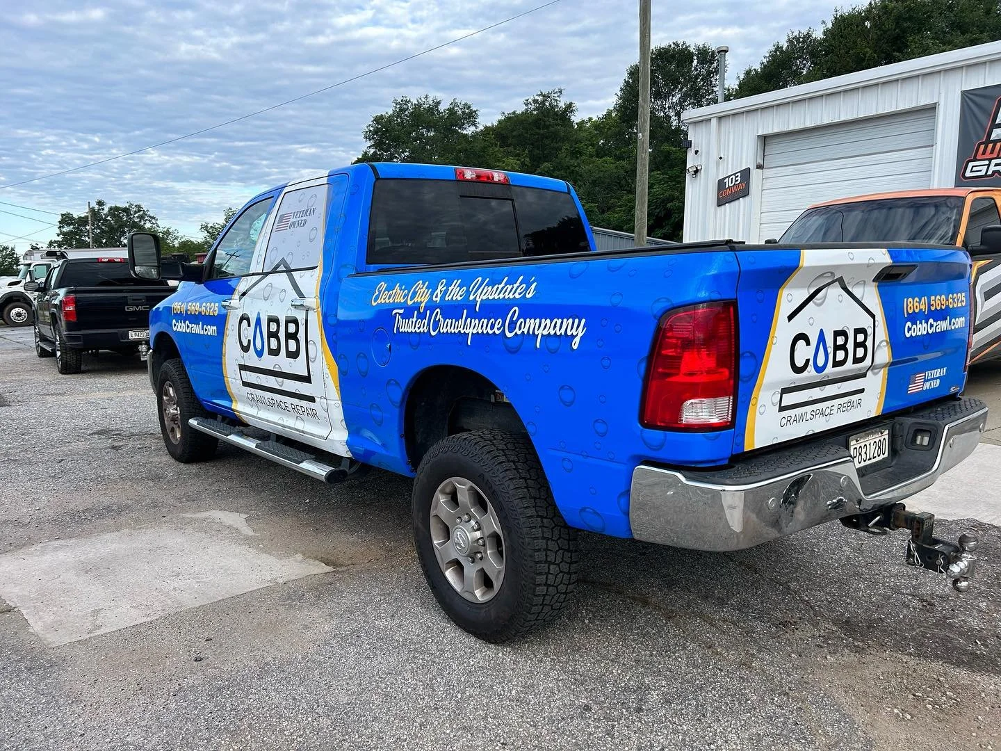 Blue pickup truck with advertising for COBB Crawlspace Repair, featuring the company logo, website, phone number, and slogan, parked on a gravel lot near a white building and other vehicles.