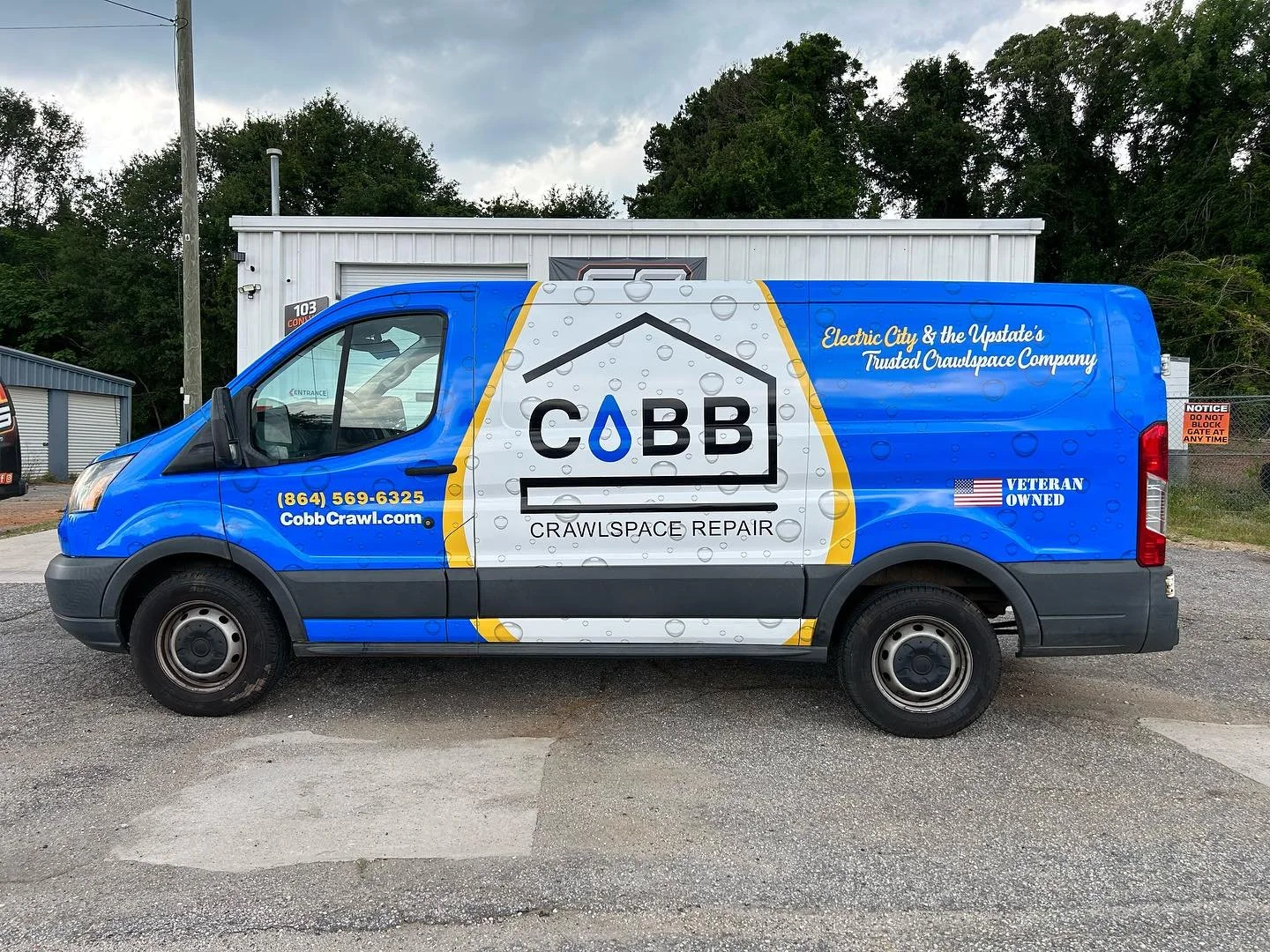 Blue service van with Cobb Crawlspace Repair branding, contact information, and slogans, parked on a gravel lot with storage units and trees in the background.