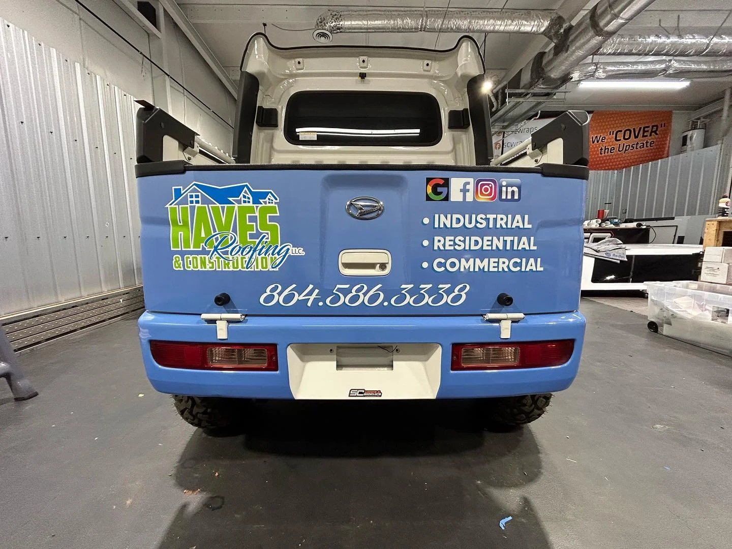 The back of a small blue utility vehicle with advertising for Haves Roofing & Construction, LLC, including contact number 864.586.3338 and social media icons for Google, Facebook, Instagram, and LinkedIn, parked inside a garage or workshop.