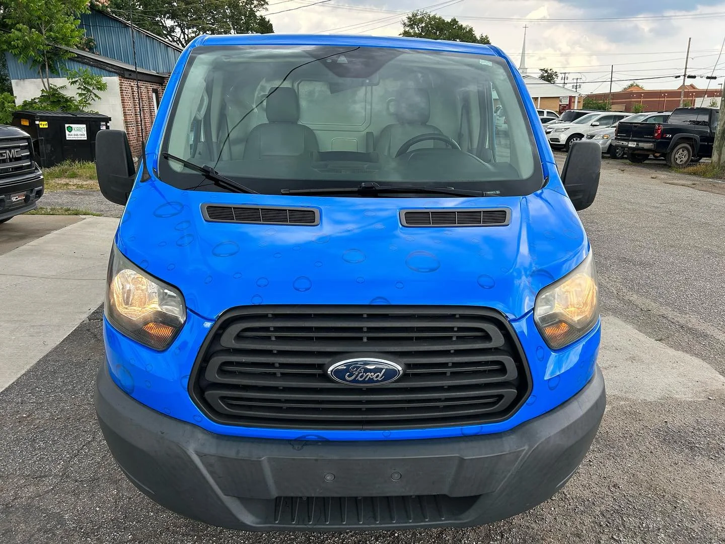 Front view of a blue Ford van parked in a lot with other vehicles in the background.