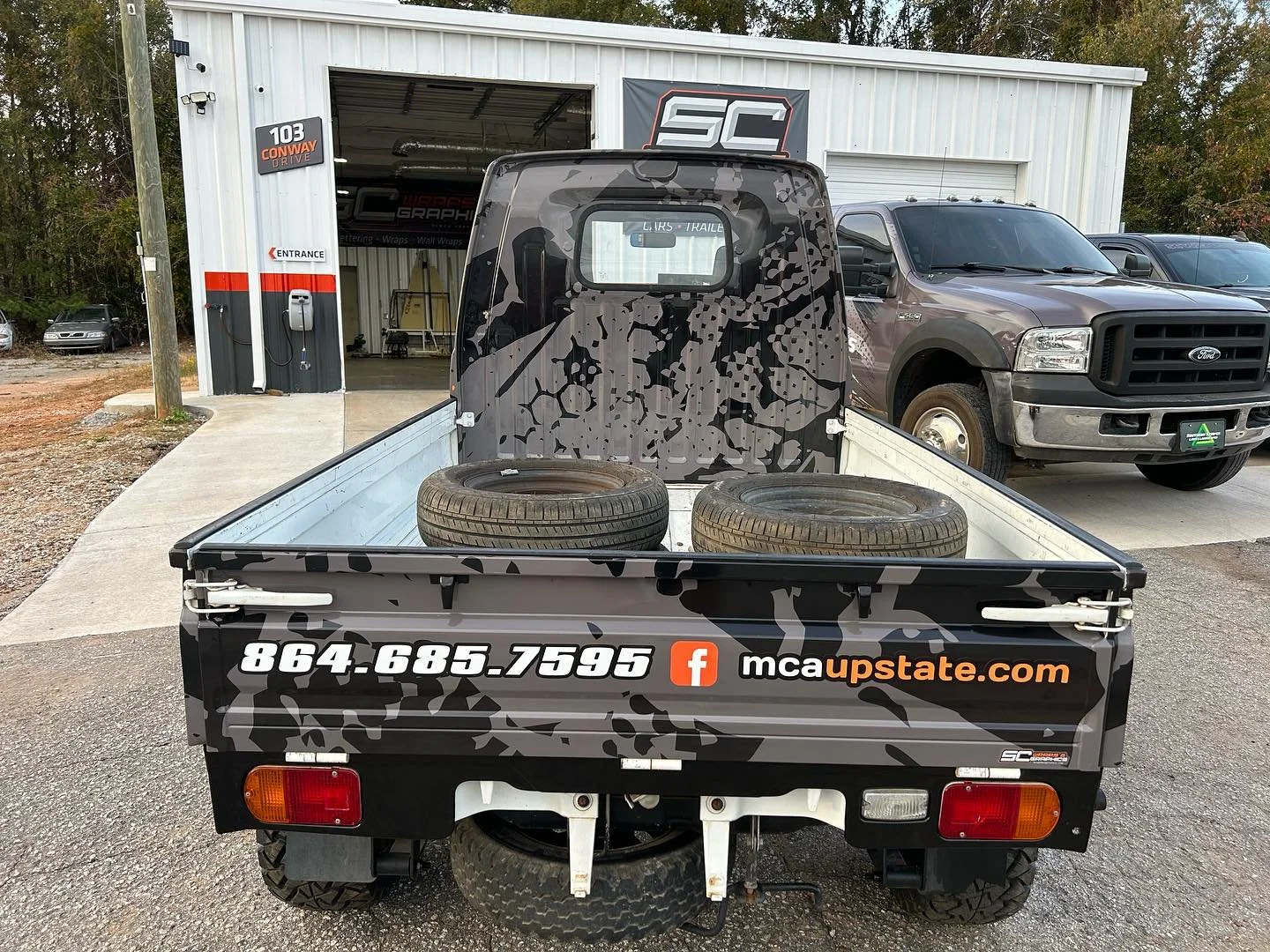 A small flatbed utility vehicle with a camouflage design and a phone number, parked outside a shop with a sign displaying 'SC' and a partially open garage door. Two tires are in the truck bed, and a larger brown pickup truck is parked beside it.