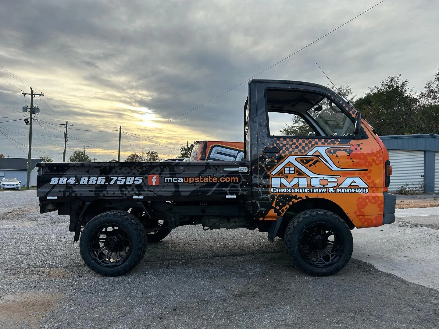 A small off-road utility vehicle with branding for MCA Construction & Roofing, featuring a black and orange color scheme, large tires, and contact information on the side, parked outdoors during sunset.
