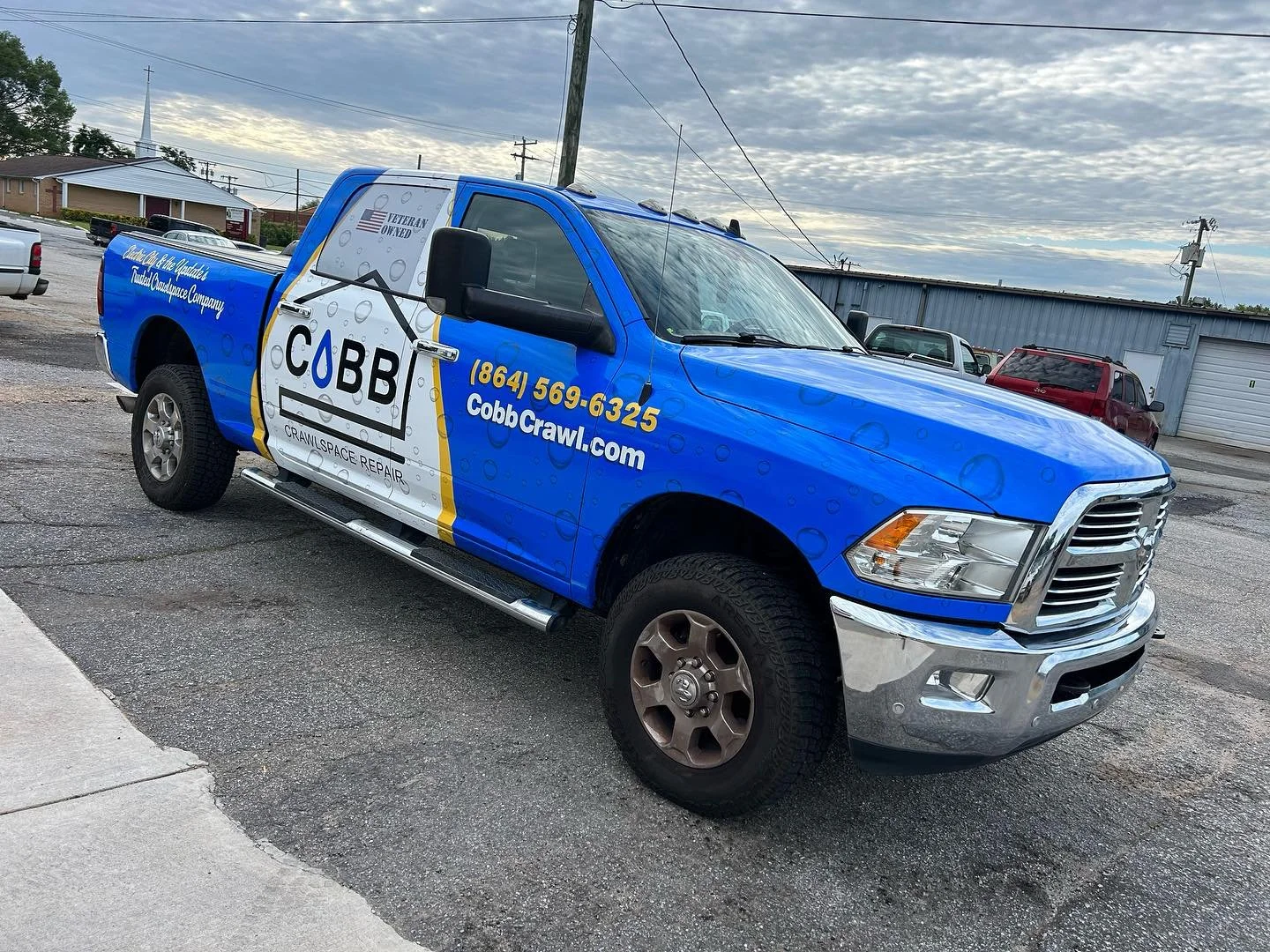 Blue pickup truck with advertising for Cobb Crawl, a crawl space repair company, parked on a street with other vehicles and a cloudy sky in the background.