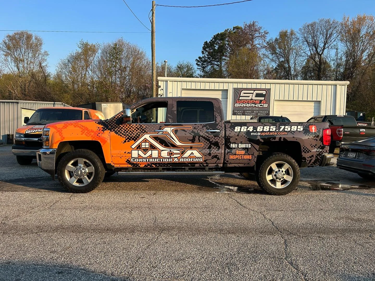 Pickup truck with advertisement for MCA Construction & Roofing parked on a gravel lot, with other vehicles and a building in the background.