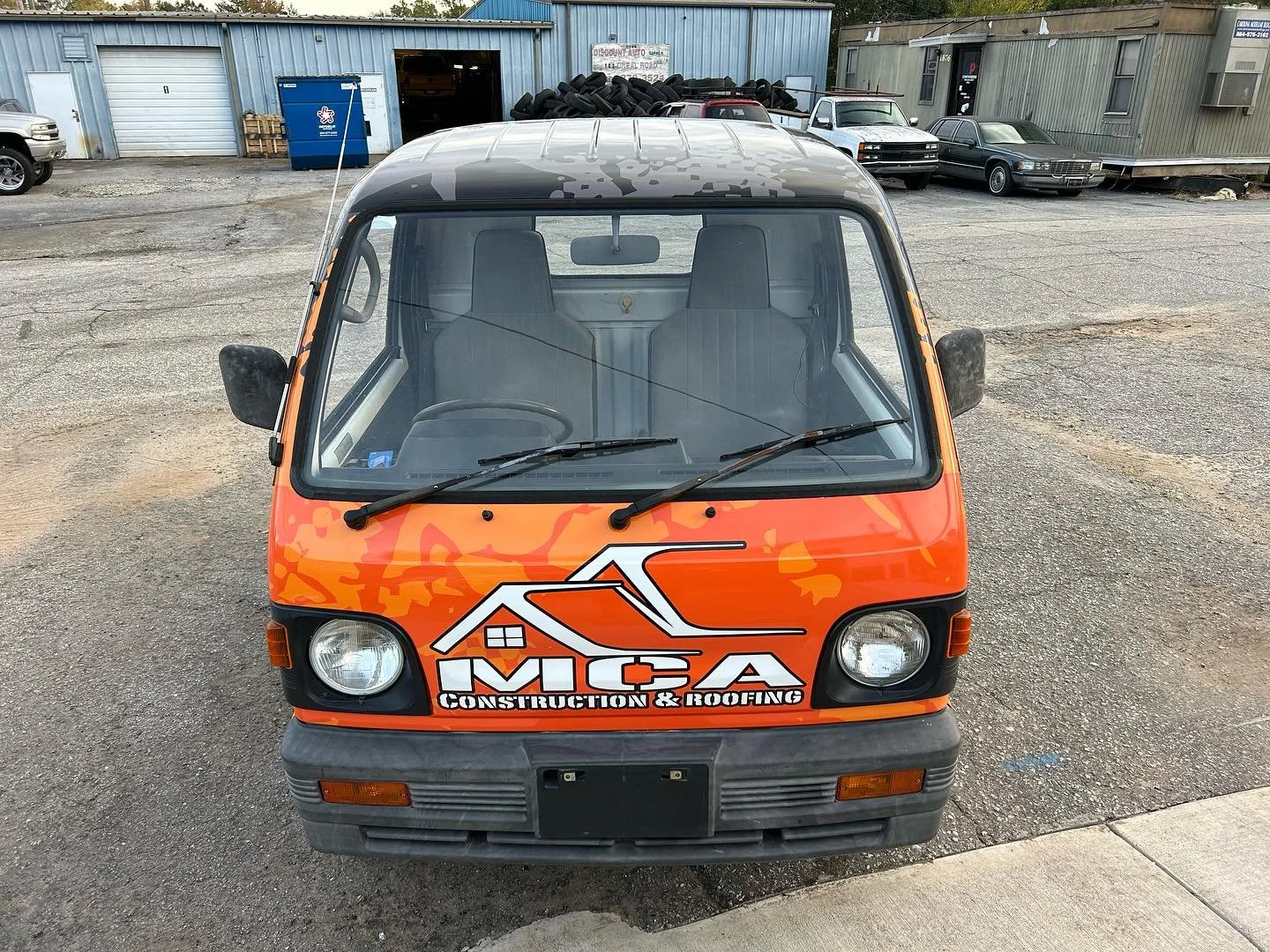 Front view of a small orange and black utility vehicle with a logo and text on the hood that reads 'MCA Construction & Roofing,' parked on a gravel surface in front of a building with tires stacked on top and other parked vehicles.