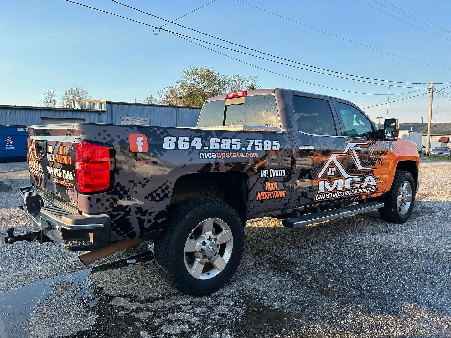 A pickup truck with advertising decals for MSA Construction & Roofing on its black and orange wrap parked on wet pavement outdoors during daylight. The vehicle features contact information, social media icons, and promotional messages for free quotes