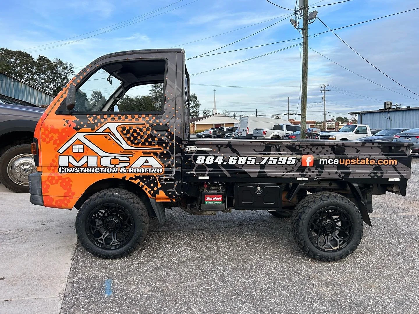 A black utility truck with orange and black graphics, displaying the name 'MCA Construction & Roofing', a phone number, website, and Facebook icon, parked on a gravel lot with other vehicles in the background.