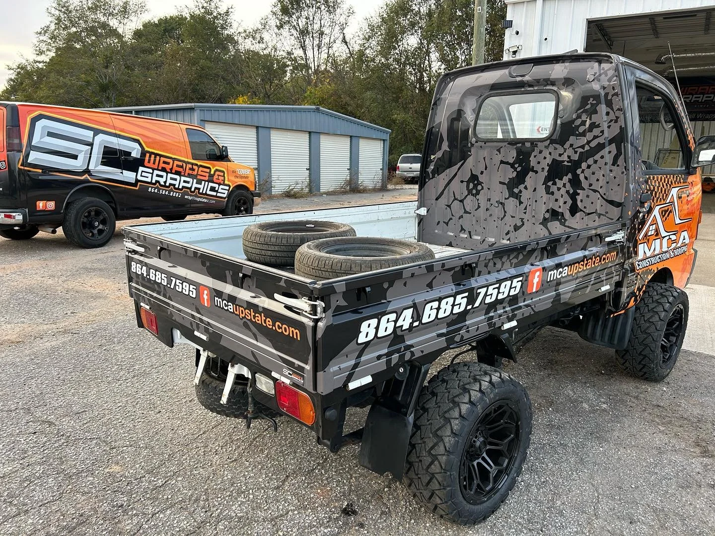 Small utility truck with a black and gray camouflage wrap promoting MCA Upstate, a construction and roofing company. The truck bed contains two spare tires and displays contact information and website.
