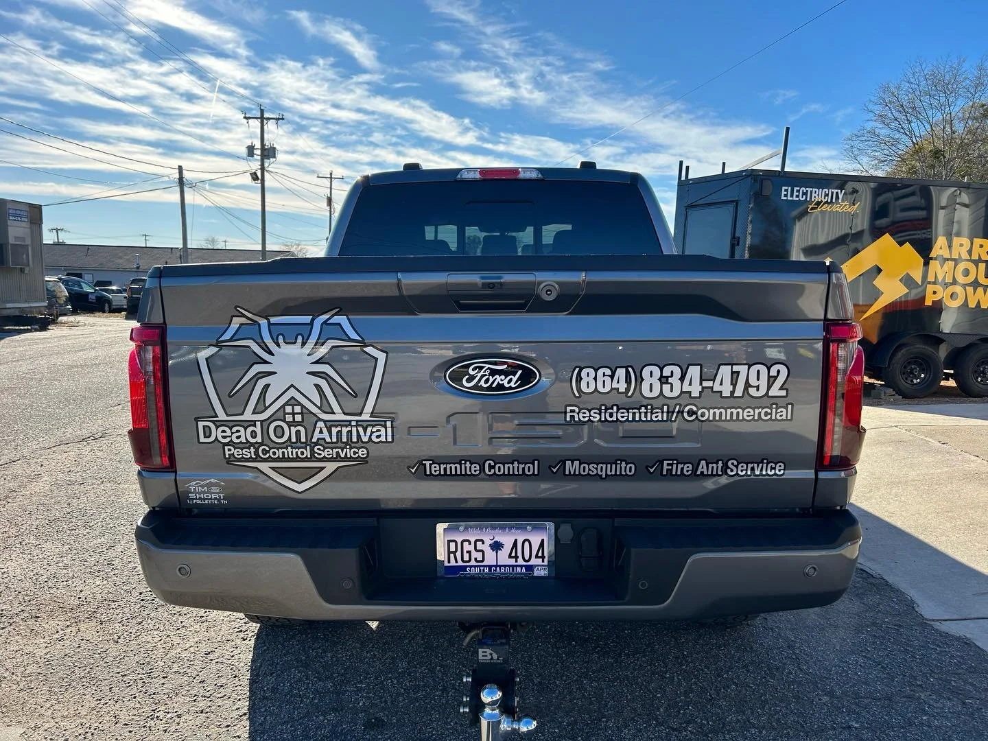 Rear view of a gray truck with pest control service advertisements, including a logo with a spider, and contact information in a parking lot with a blue sky and a few clouds.