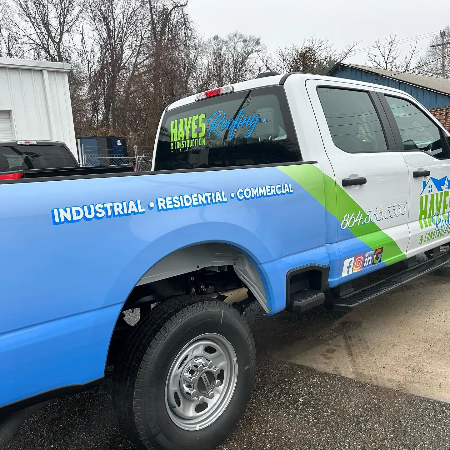 Company truck for Hayes Roofing & Construction with blue, green, and white graphics displaying contact information and services, parked outdoors in front of a building and trees.