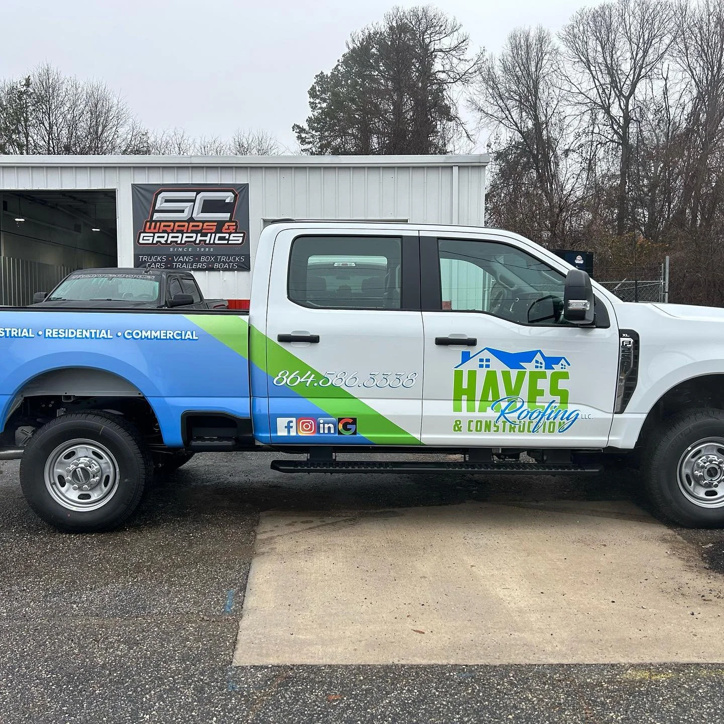A company truck for Hayes Roofing & Construction parked outside a building with a banner for SC Wraps & Graphics in the background. The truck displays contact information and services, with a partially cloudy sky and leafless trees behind.