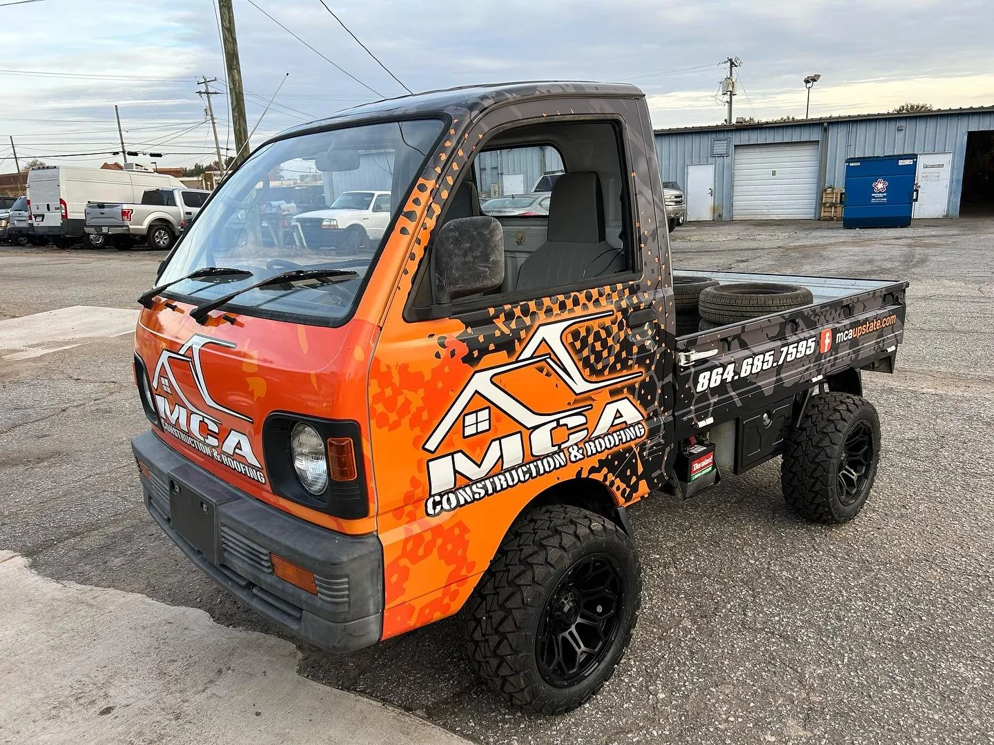 Small utility truck with WCA Construction & Roofing logo, orange and black wrap, large off-road tires, parked on asphalt with other vehicles and a building in the background.