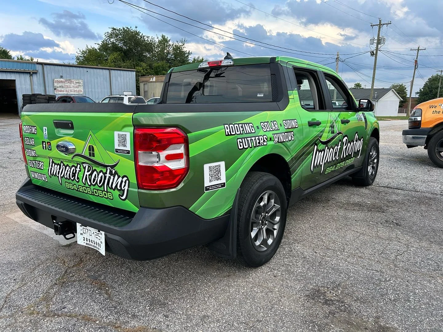 Green Impact Roofing pickup truck with the company's logo and contact information parked on a gravel lot with a blue building and other vehicles in the background.