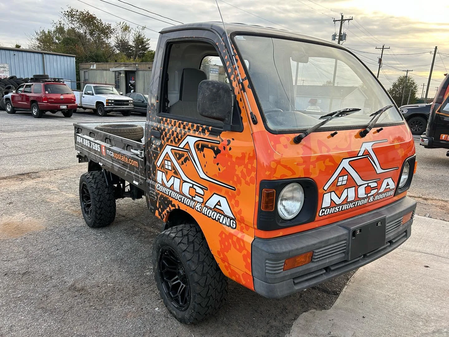 An orange mini truck with black off-road tires and construction company branding, including the logo and website for MCA Construction & Roofing, parked in a lot with other cars and vehicles.