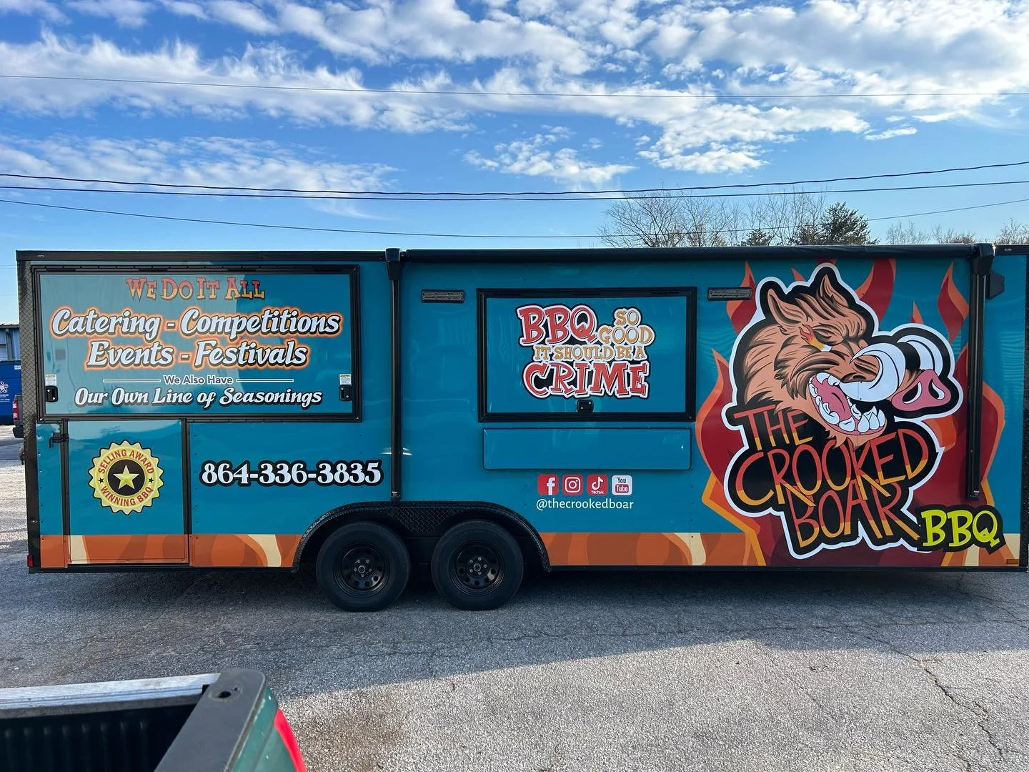 Blue food truck named The Crooked Boar BBQ with colorful graphics, a smiling boar cartoon mascot, and text advertising catering, competitions, events, festivals, seasoned barbecue, and a humorous slogan about BBQ being so good it should be a crime. The truck displays social media icons and a phone number.