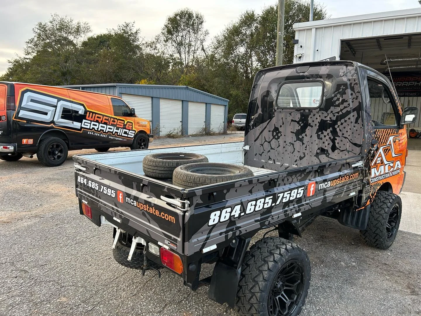 A black utility vehicle with branding for MCC Upstate, parked outdoors on a gravel lot, with two tires in the cargo area, and a similar branded van in the background, near a blue building with garage doors and trees.