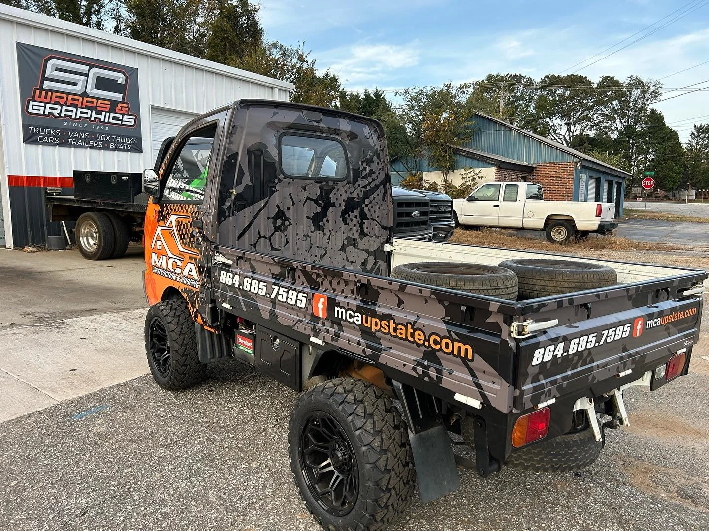 Colorful utility truck with graphics, advertisement for MCA Construction & Roofing, parked outside a building with signage for SC Wraps & Graphics, and other vehicles in the background.