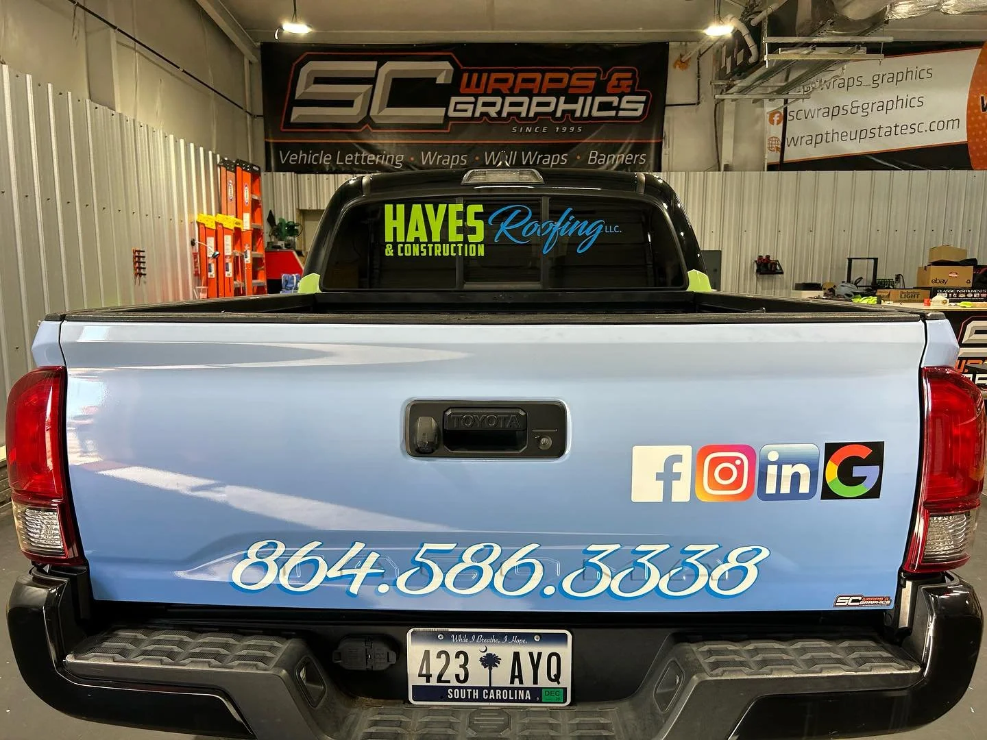 The back of a light blue pickup truck with a South Carolina license plate reading '423 AYO.' The truck has various decals, including social media icons and a phone number. The decals include Facebook, Instagram, LinkedIn, and Google. There is also a 