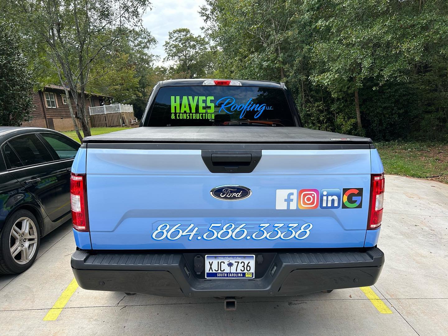 Rear view of a blue Ford pickup truck with company name, phone number, social media icons, and a decal for Hayes Roofing and Construction, parked in a lot with a black vehicle on the side and green trees in the background.