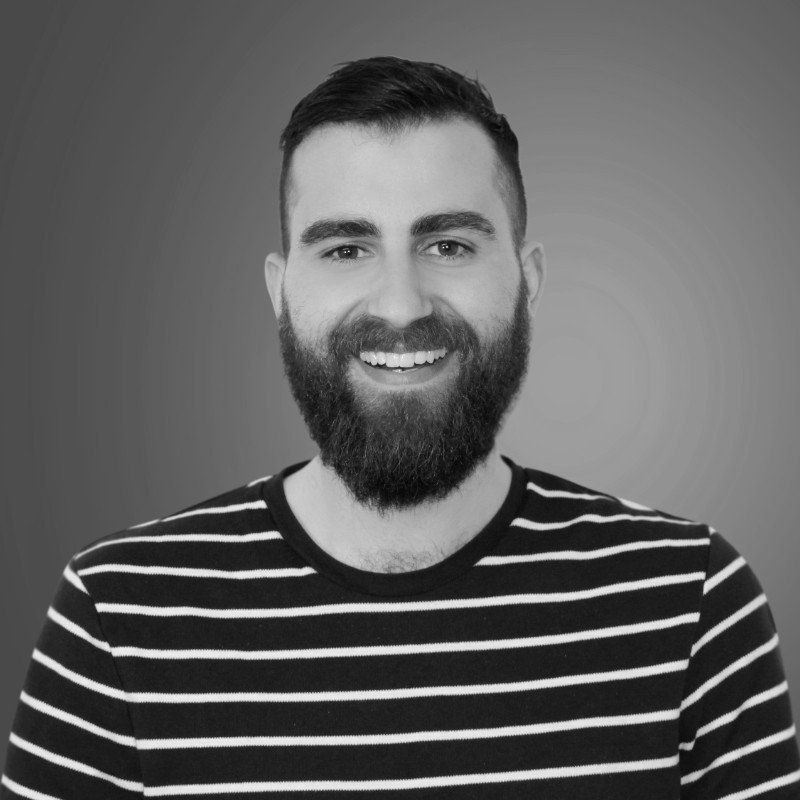 A smiling man with a beard and short dark hair, wearing a striped shirt, standing against a plain background.