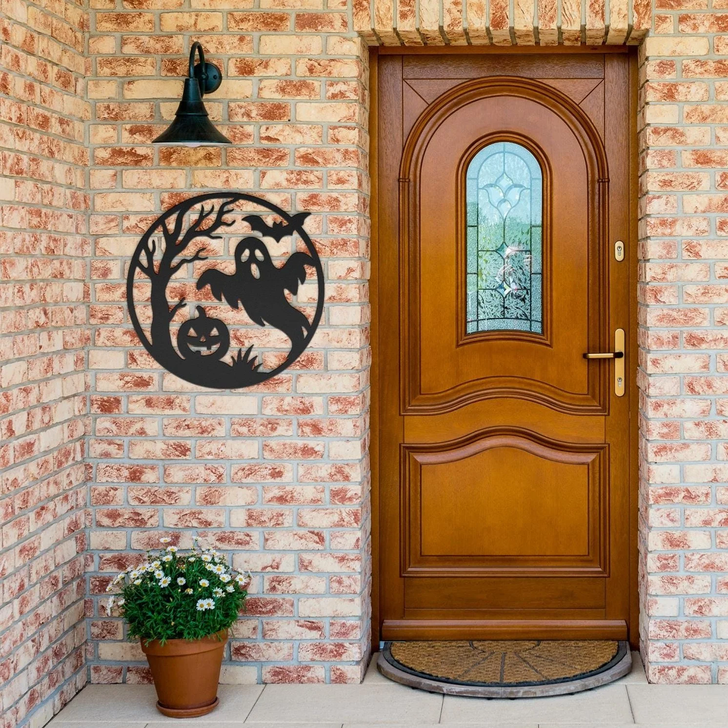 Front door with Halloween decoration: a black metal ghost and pumpkin with a face, hanging on a brick wall next to a flowerpot with white daisies.