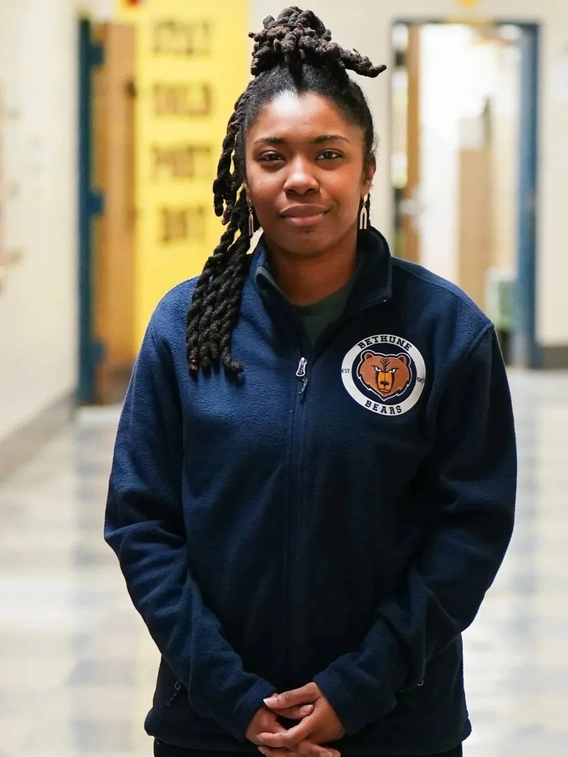 A woman with braided hair standing in a school hallway, wearing a navy jacket with a 'Bethune Bears' logo.