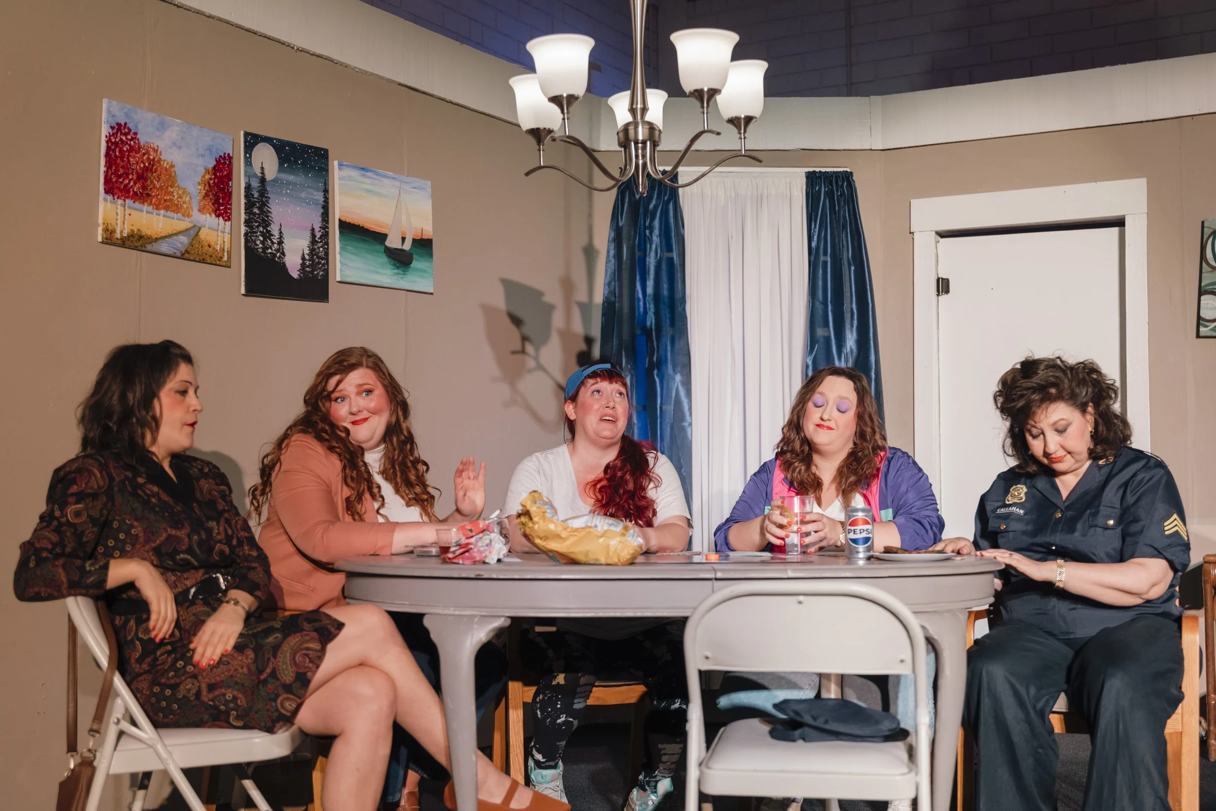 Five women sitting around a dining table, engaging in conversation. The women show various facial expressions, some smiling and others with serious looks. There are food and drink items on the table, and a woman in a police uniform is present. The room has beige walls with three landscape paintings, blue curtains covering a window, and a ceiling chandelier with seven lights.