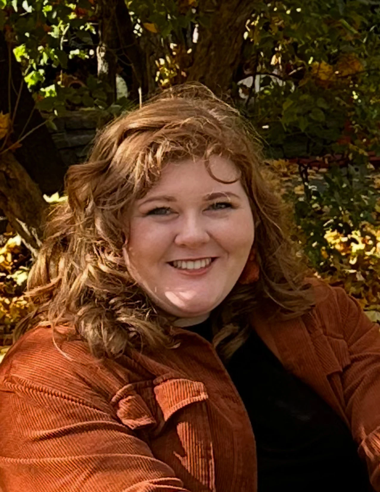 A woman with curly red hair smiling outdoors with autumn leaves in the background.