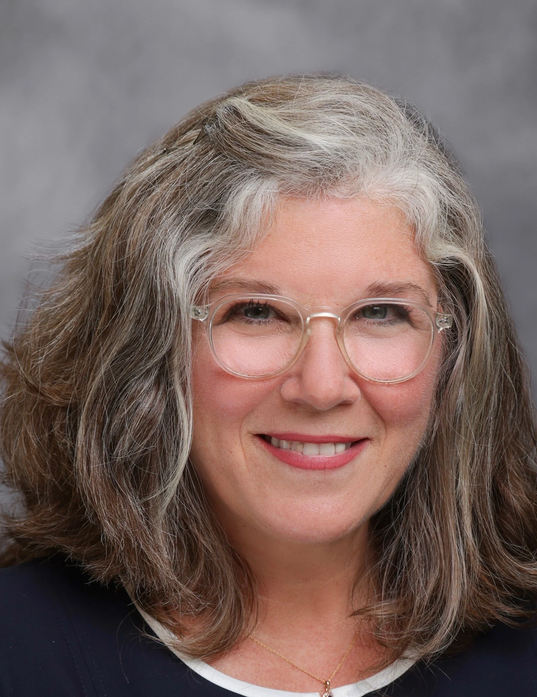 Close-up of a smiling woman with gray, curly hair and glasses, wearing a navy blue top and a gold necklace, against a gray background.