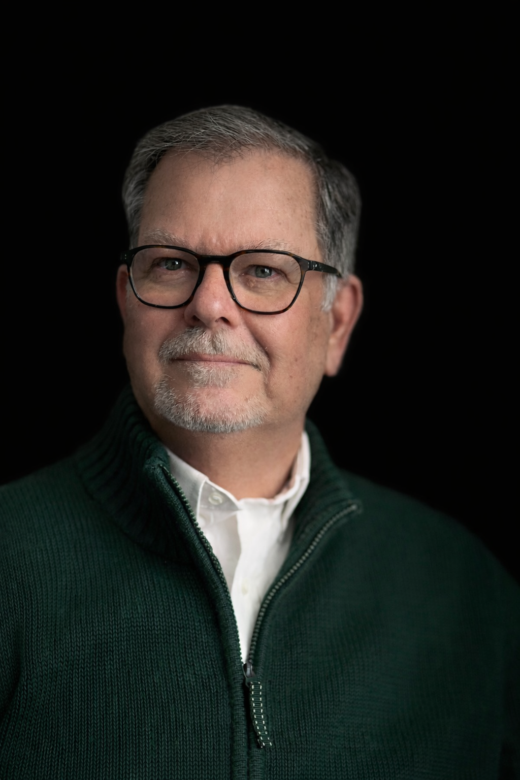 Close-up portrait of a middle-aged man with gray hair, glasses, and a gray beard, wearing a white shirt and dark green zip-up sweater, against a black background.