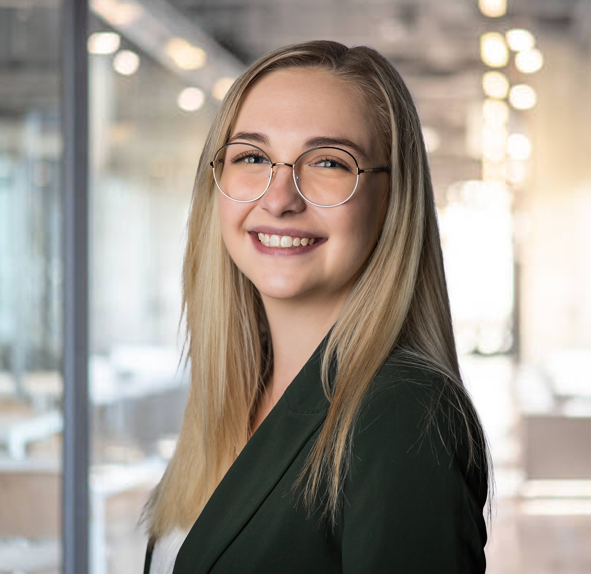 Close-up of a smiling woman with blonde hair, wearing glasses and a black blazer, in an indoor setting with blurred background lighting.