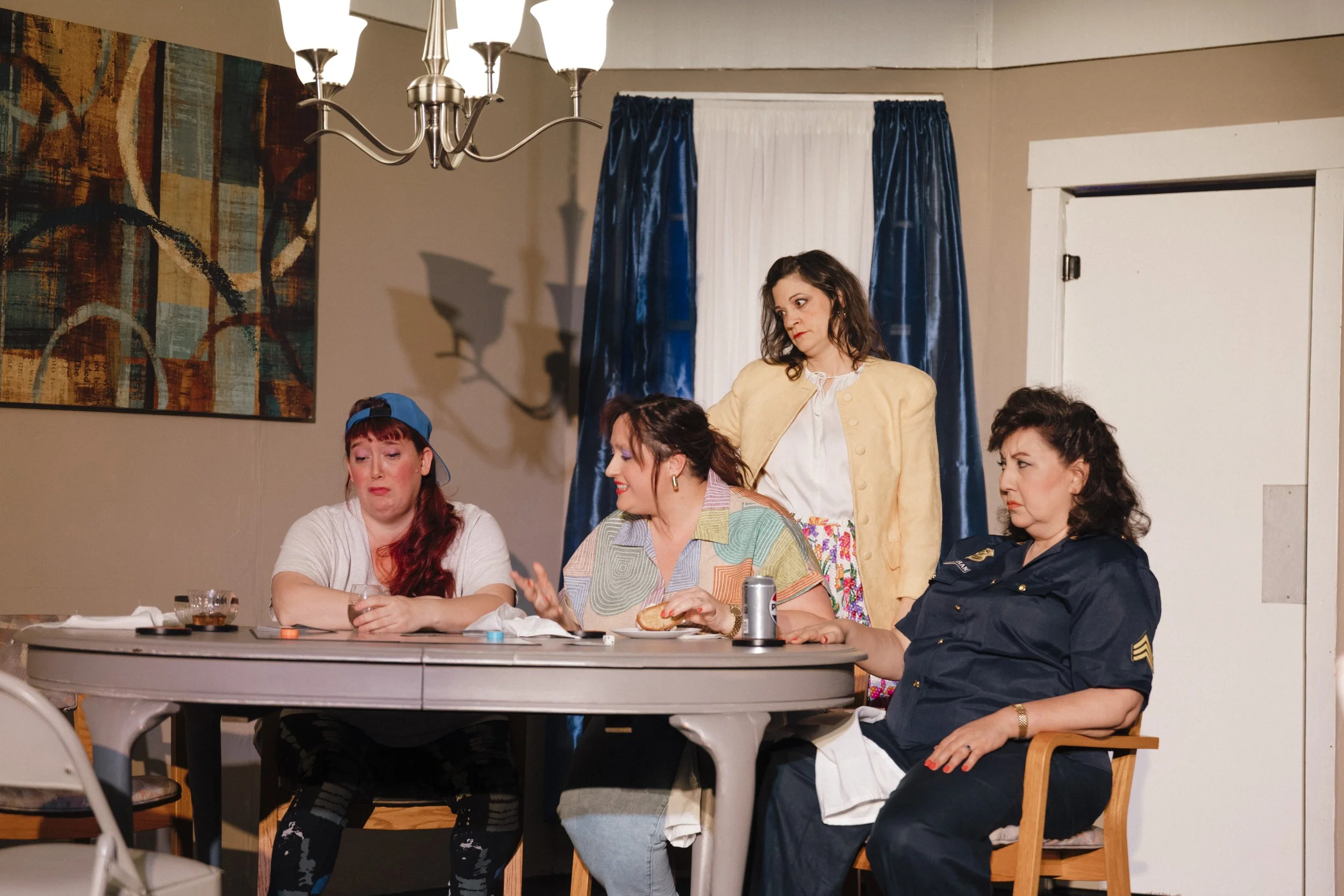 A group of five women sitting at a round table with drinks and snacks, engaged in conversation, with one standing woman looking over them, in a room with abstract art on the wall and blue curtains.