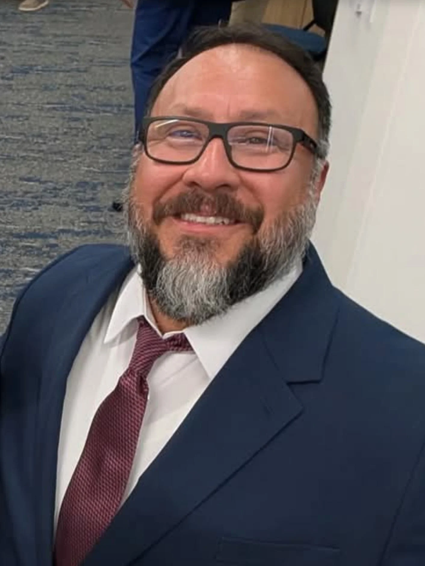 A smiling man with glasses, a beard, and dark hair, dressed in a navy suit, white shirt, and maroon tie, at an indoor professional setting.