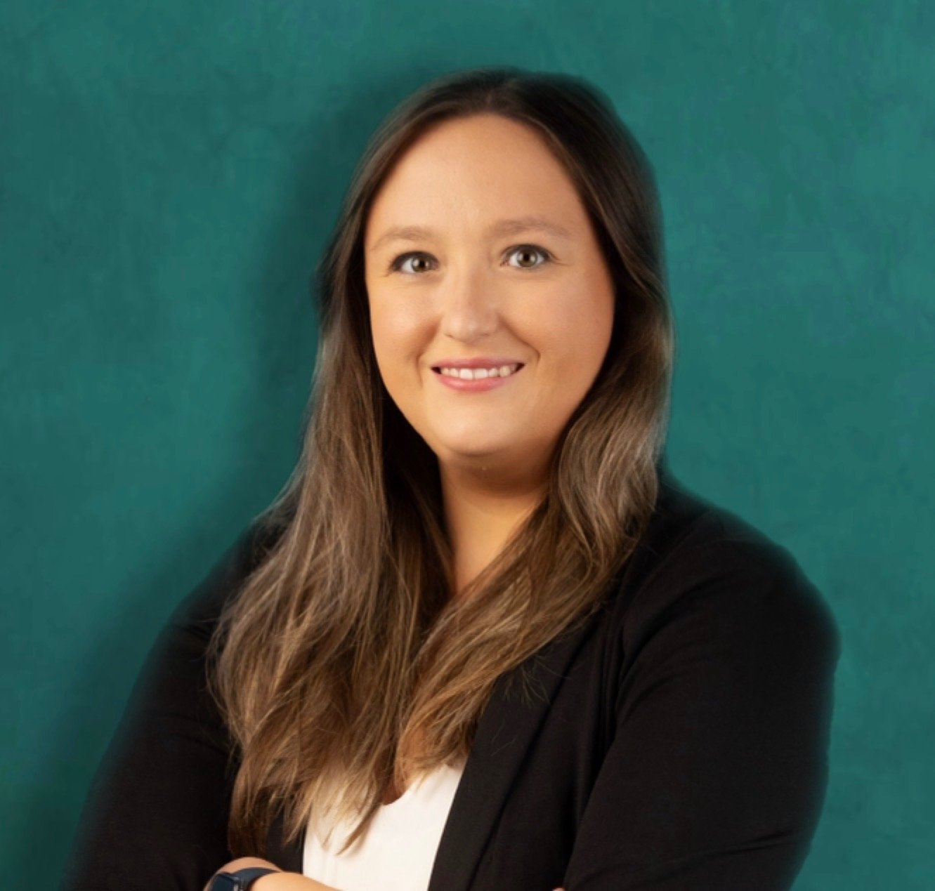 A woman with long brown hair smiling, wearing a black blazer and white shirt, standing against a green background.