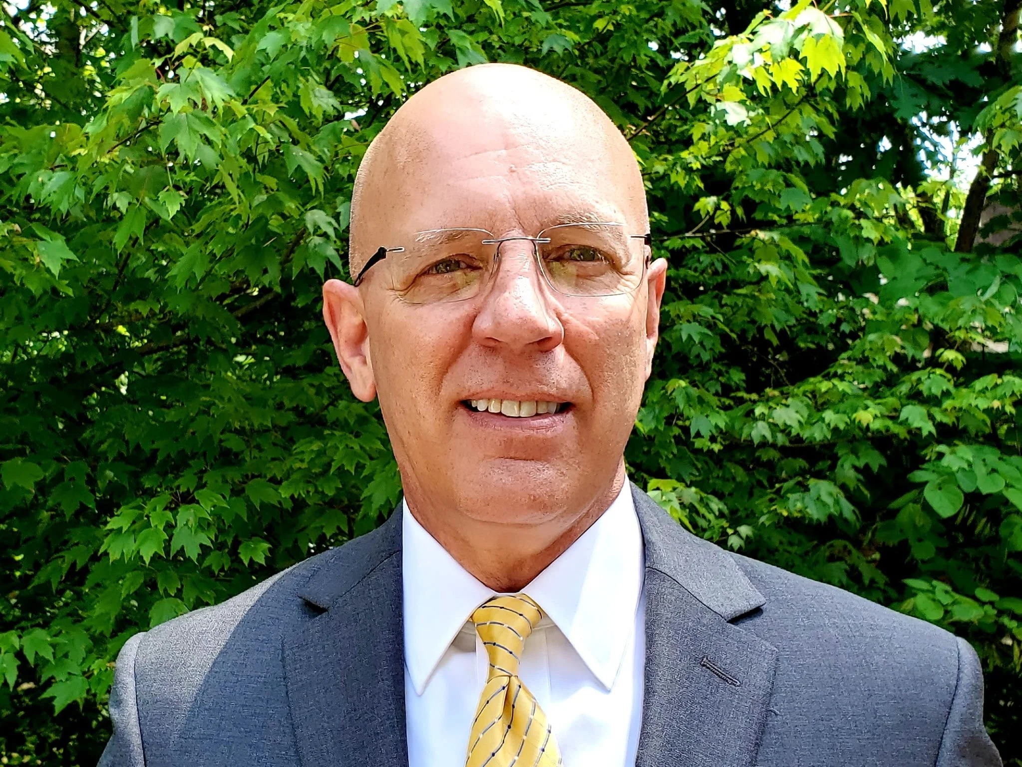 Portrait of a smiling bald man with glasses, wearing a gray suit jacket, white shirt, and yellow striped tie, standing in front of leafy green trees.