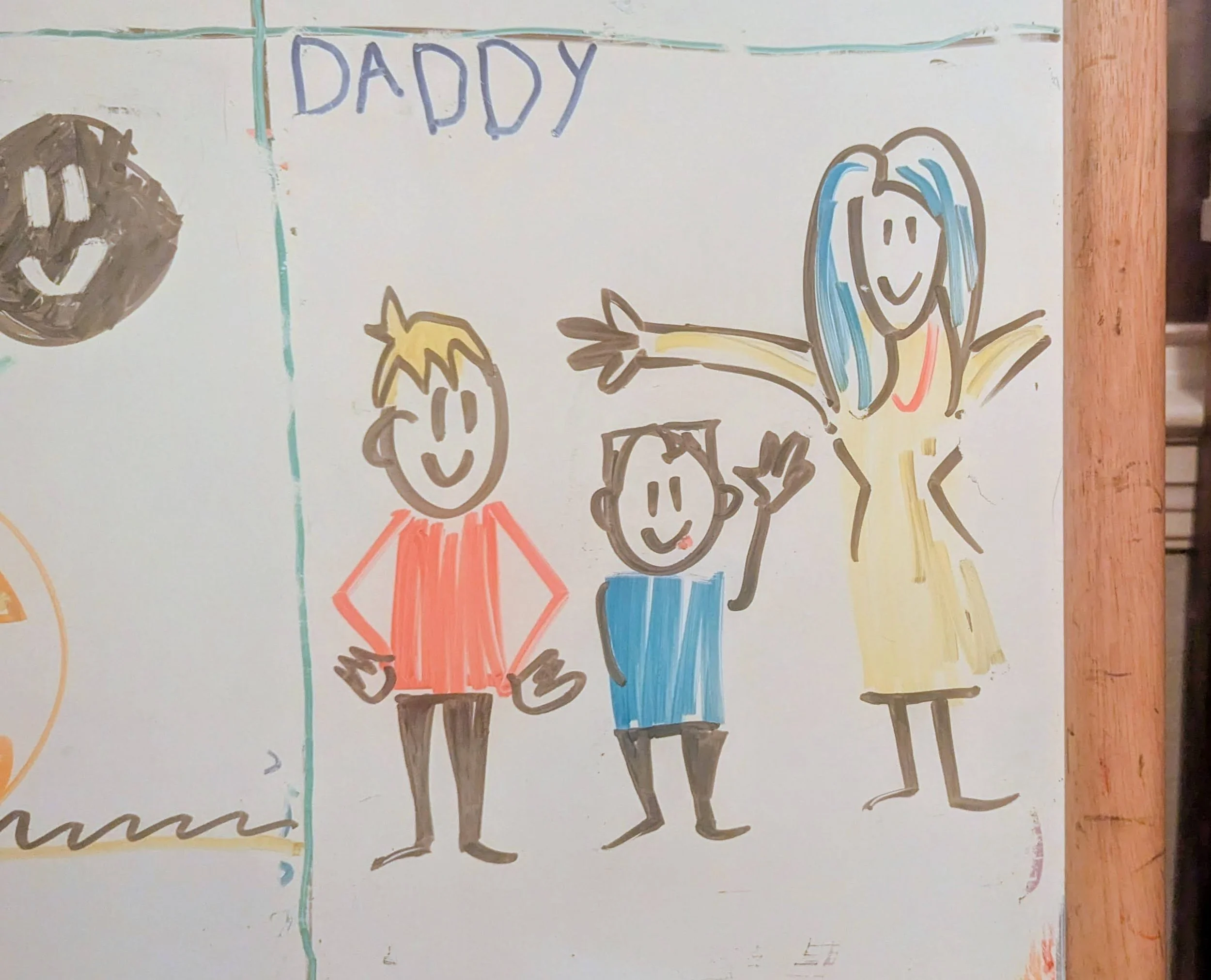 Colorful drawing of three happy people, with the word 'DADDY' written at the top. The people include a woman with blue hair wearing a yellow dress, a boy in a blue shirt, and a man in a red shirt. They are smiling and posing cheerfully.
