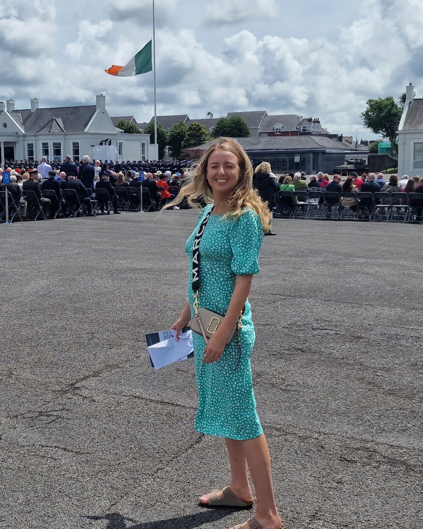 A woman in a turquoise polka dot dress smiling outdoors at a large gathering with many seated people, a flag flying, and buildings in the background.