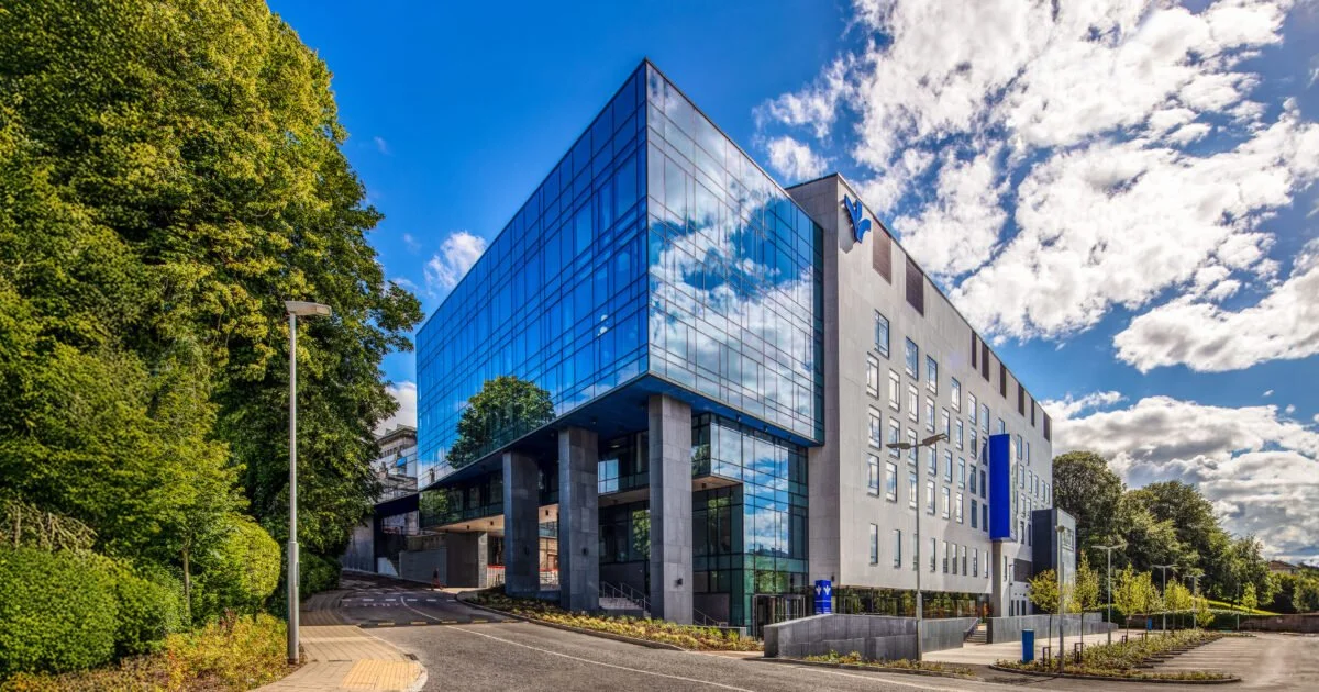Modern multi-story hospital building with glass facade reflecting the blue sky and clouds, with green trees on the left and a clear road in front.