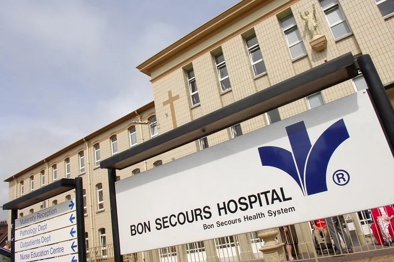 Sign for Bon Secours Hospital with directions to maternity reception, pathology department, outpatients department, and nurse education centre outside a hospital building.