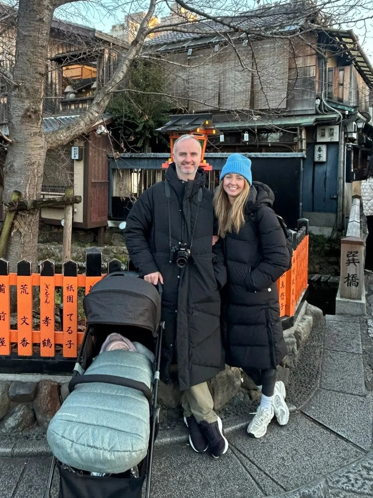 A man and woman smiling while standing outdoors, with the man holding a camera and pushing a stroller with a sleeping child, in front of a traditional Japanese building decorated with orange and black signs with Japanese writing.