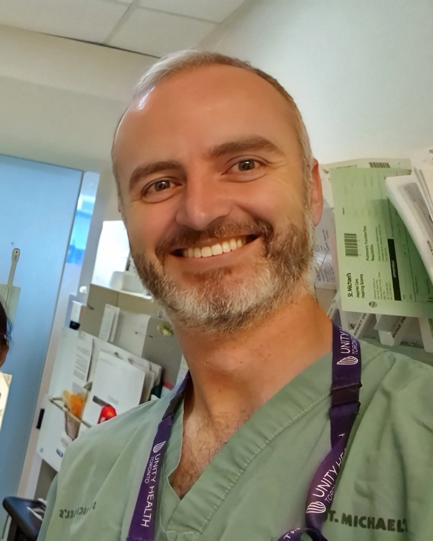 A smiling man wearing green medical scrubs and a lanyard in a hospital or clinic setting with medical charts and equipment in the background.