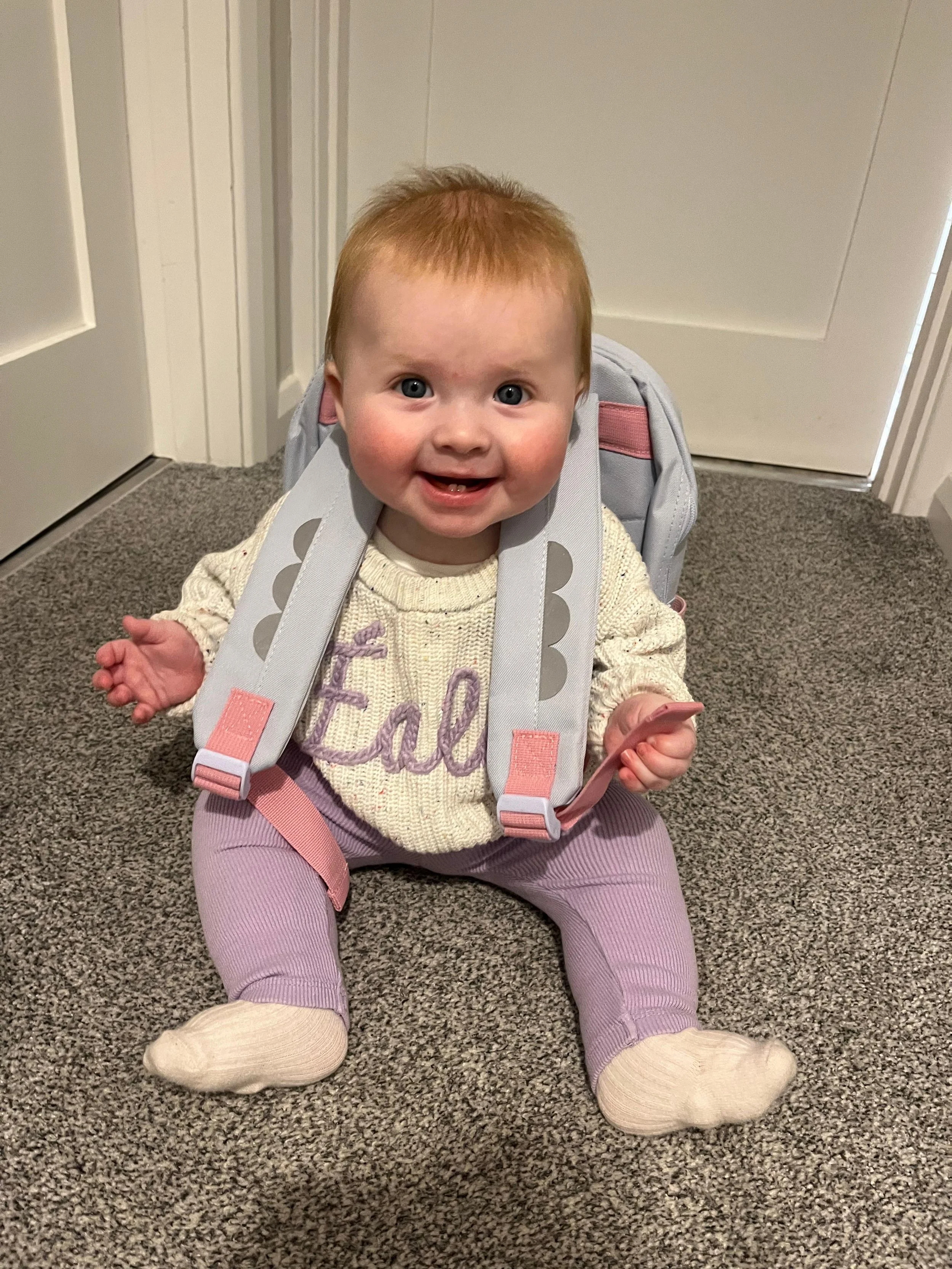 A smiling baby girl with red hair and blue eyes sitting on a carpeted floor, wearing a cream sweater, pink pants, cream socks, and a gray and pink backpack.