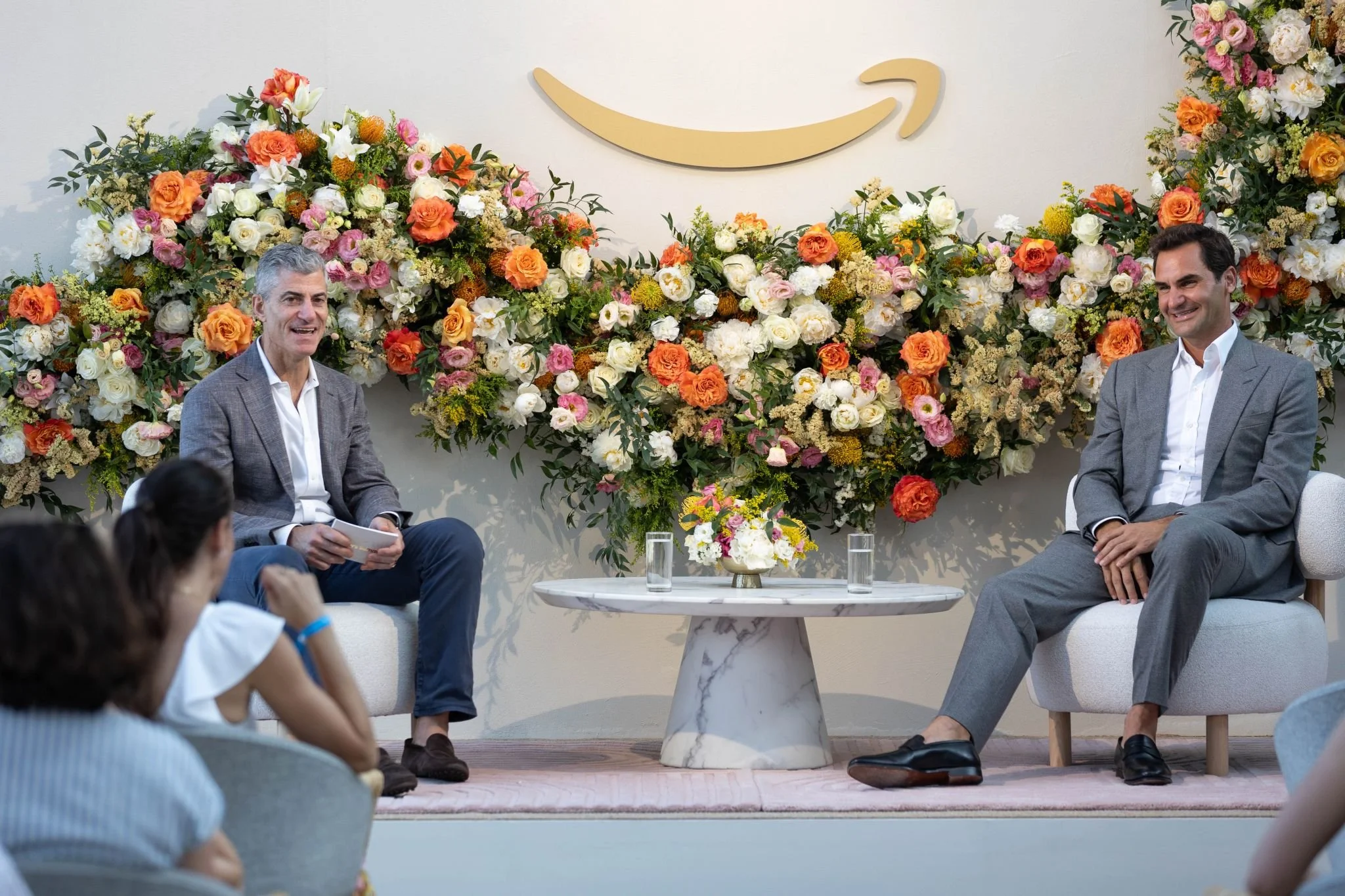 Two men in suits sit on a stage decorated with pink, white, and orange flowers, with a large Amazon smile logo above them. They are having a conversation while a small group of people listens.