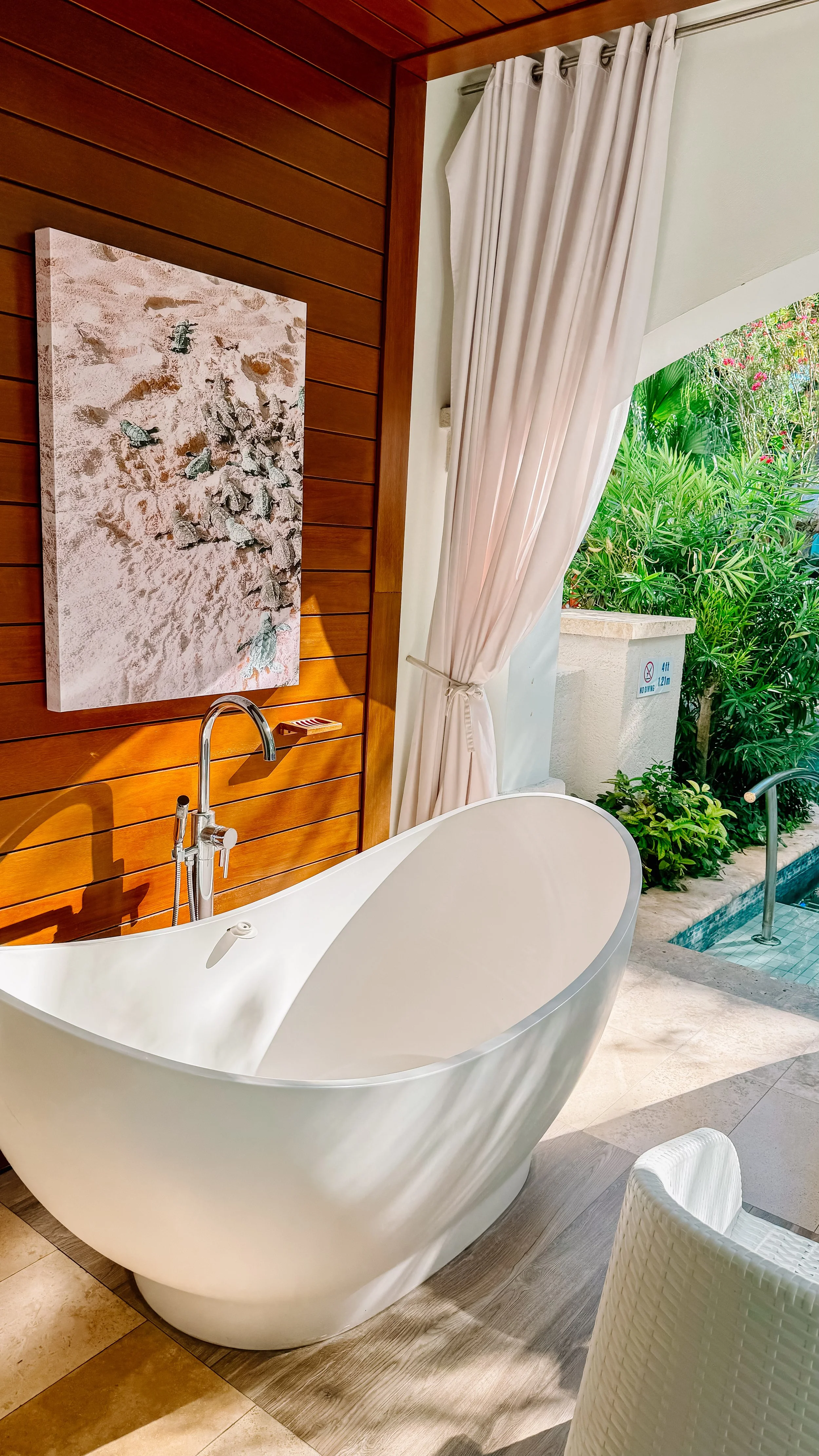 A modern bathroom with a freestanding bathtub, wooden wall paneling, framed artwork, and an outdoor view of greenery.