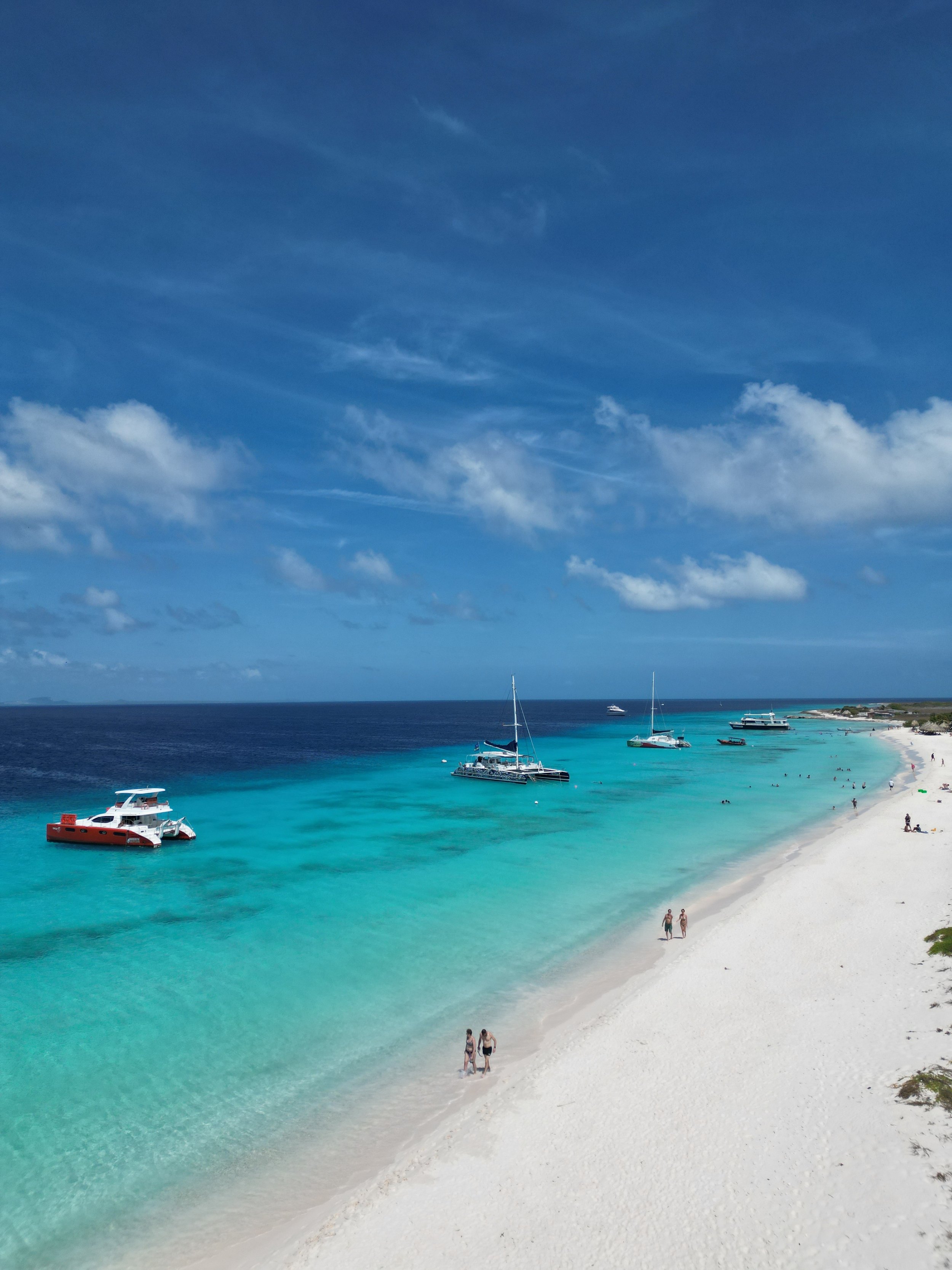 Clear blue sky with wispy clouds, turquoise water with boats, and white sandy beach with a few people walking and relaxing.