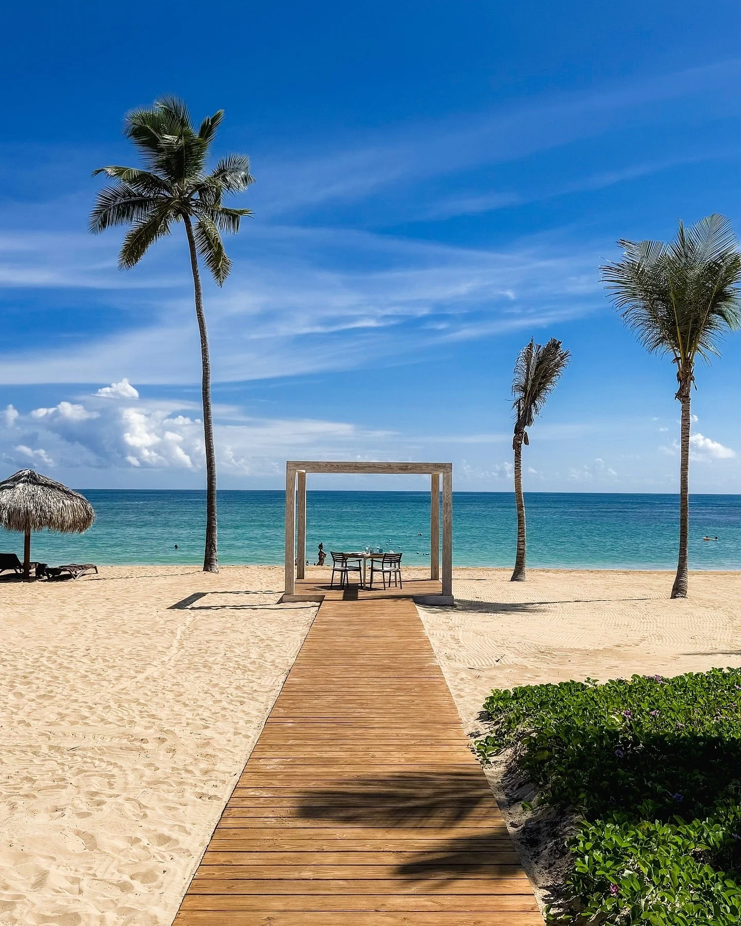 A tropical beach with a wooden walkway leading to a picnic table under a modern wooden frame, with tall palm trees, a thatched umbrella, and the ocean in the background under a blue sky with some clouds.