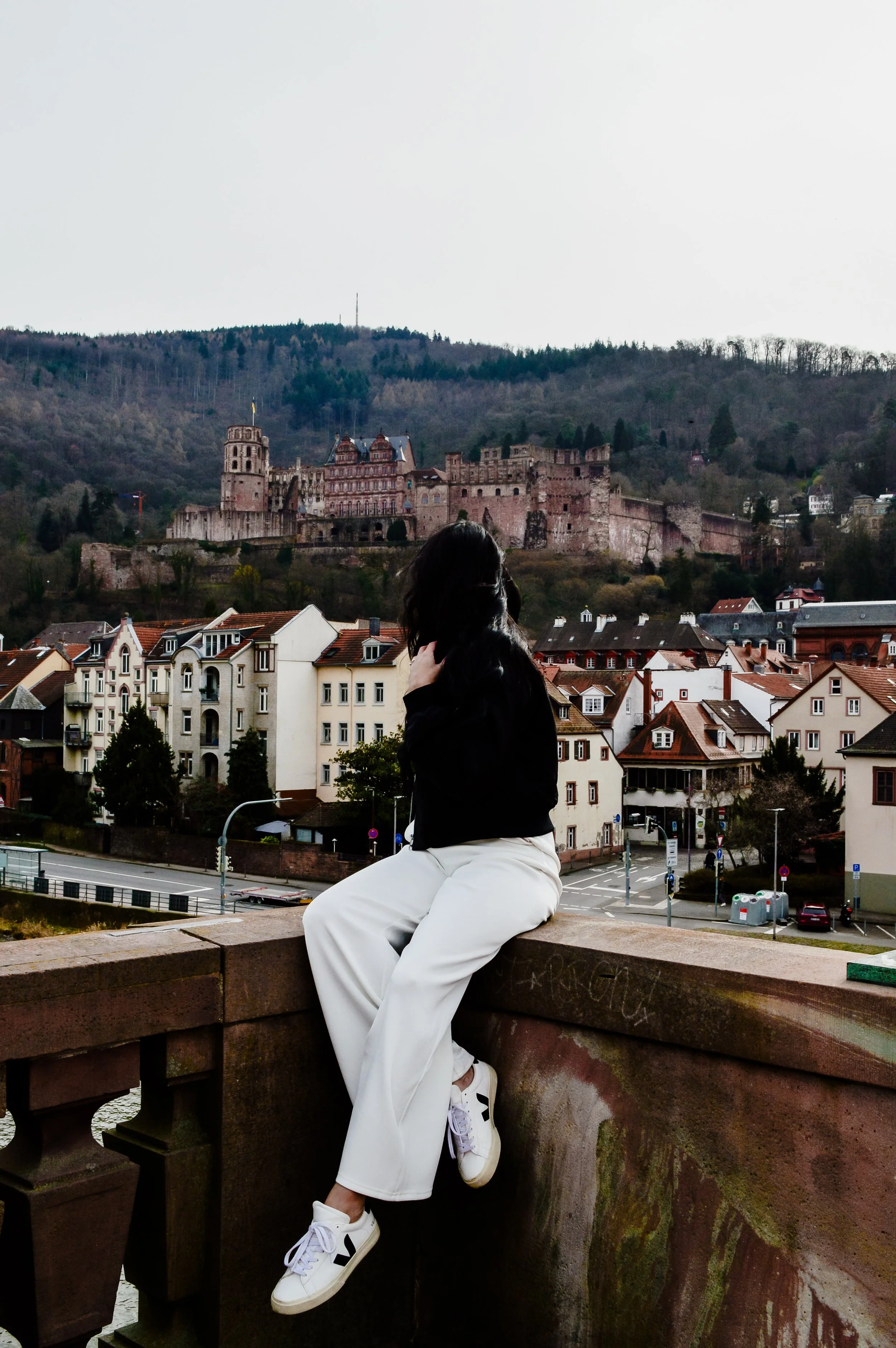 A woman sitting on a bridge ledge overlooking a European town with a castle on a hillside in the background.