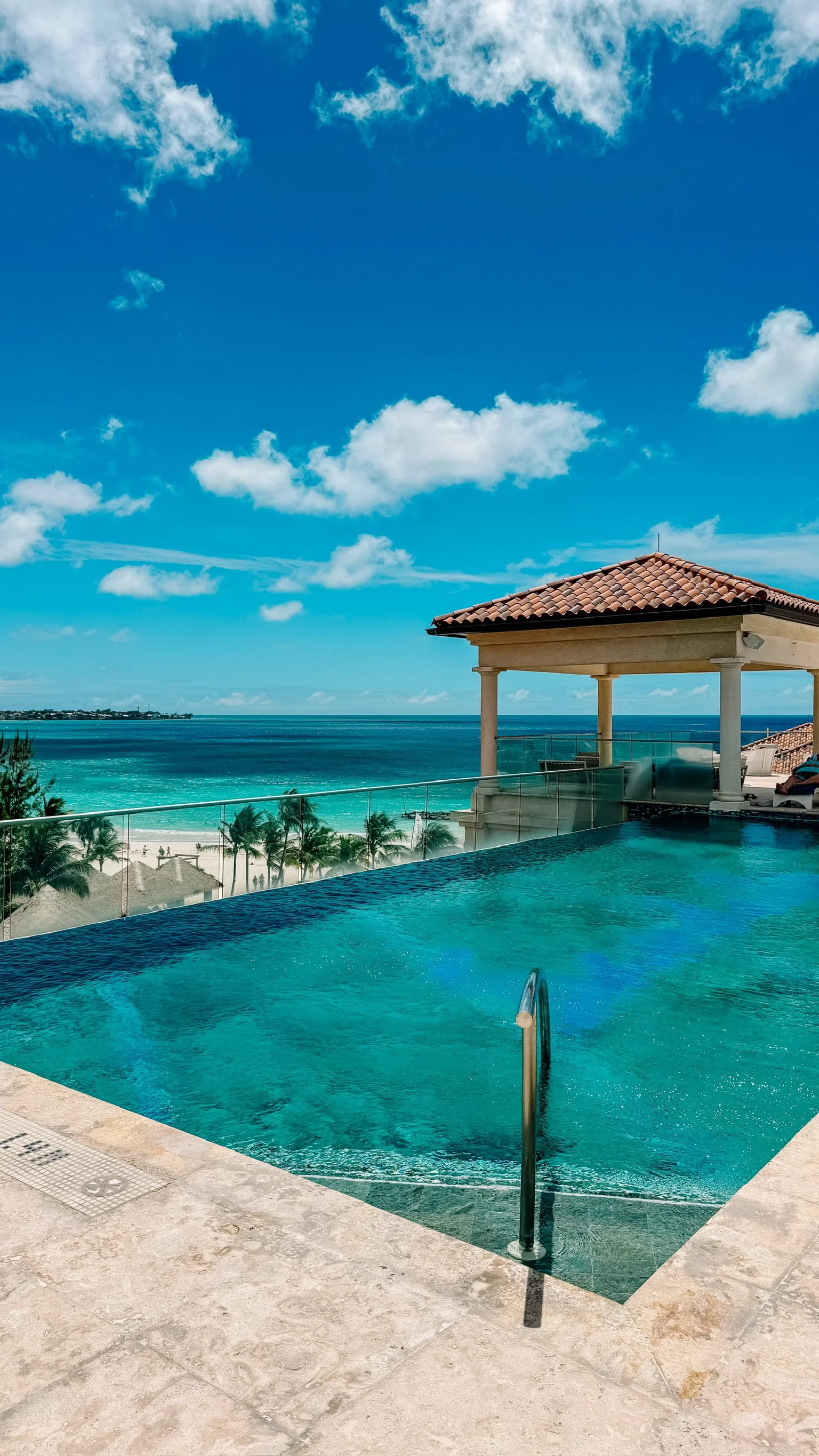 An infinity pool overlooking the ocean, with a deck and chairs under a tiled-roof pavilion, palm trees, and a clear blue sky with scattered clouds.