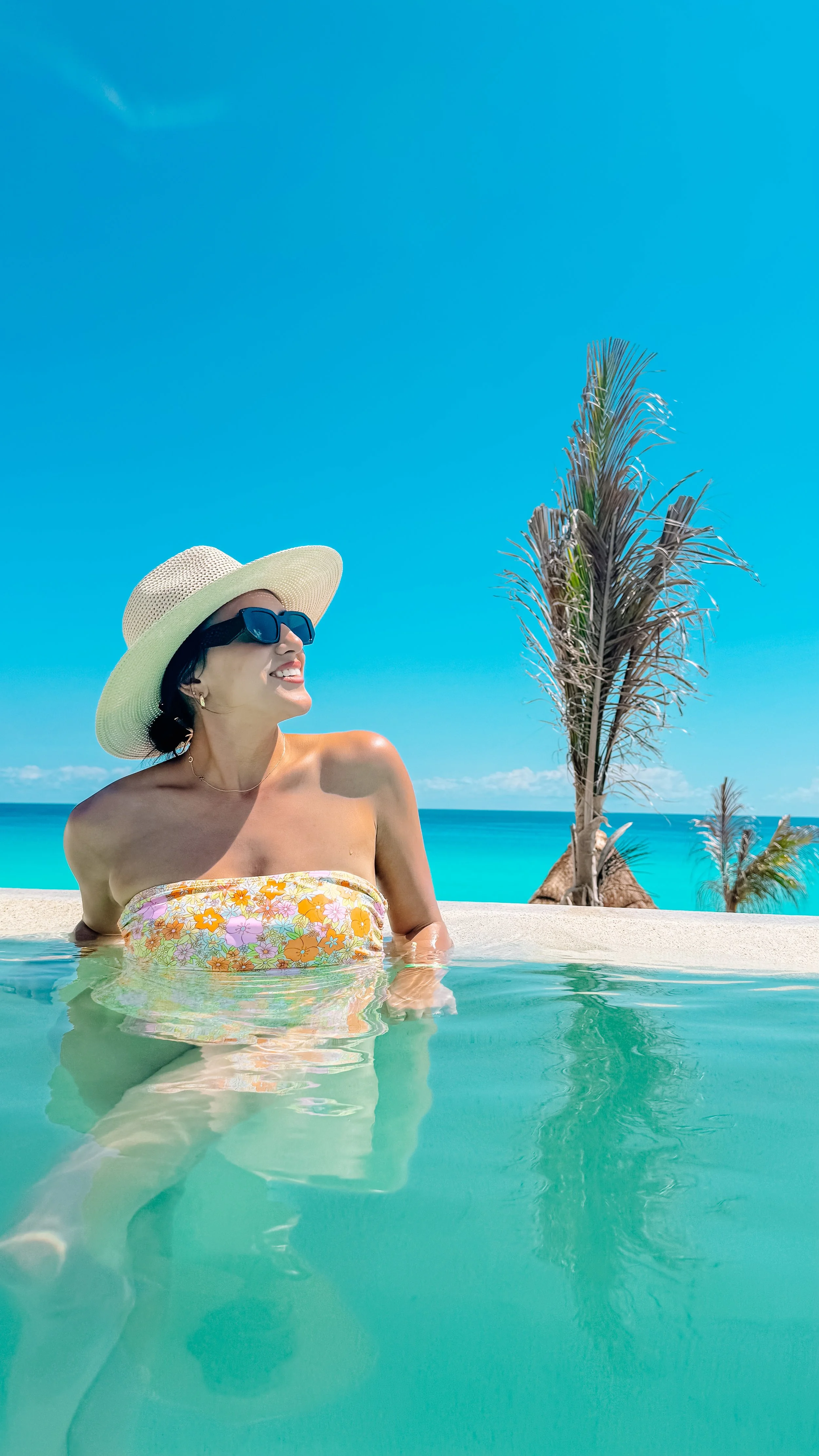 A woman wearing sunglasses, a wide-brimmed hat, and a floral strapless swimsuit, sitting in a swimming pool with a tropical beach background, including palm trees and blue sky.